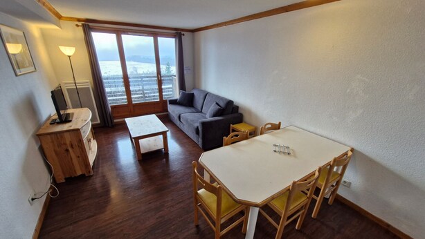 A small, cozy room with a wooden desk, a gray sofa, a dining table with chairs, and a large window offering a view of a snowy landscape.