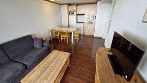 A compact living space with a sofa, a wooden coffee table, a TV on a wooden stand, a dining table with chairs, and a kitchenette in the background.