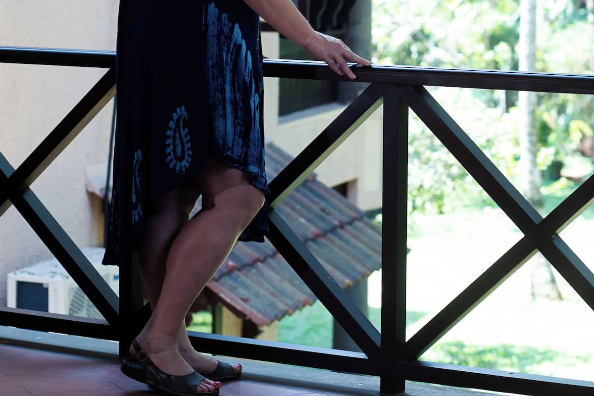 A person standing on a balcony with a view of greenery and a building with a tiled roof.