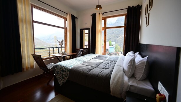 A bedroom with a large bed, a desk, and a chair, a window with a view of mountains, and a ceiling light. - The Bulevar - Alpine (Lahul)