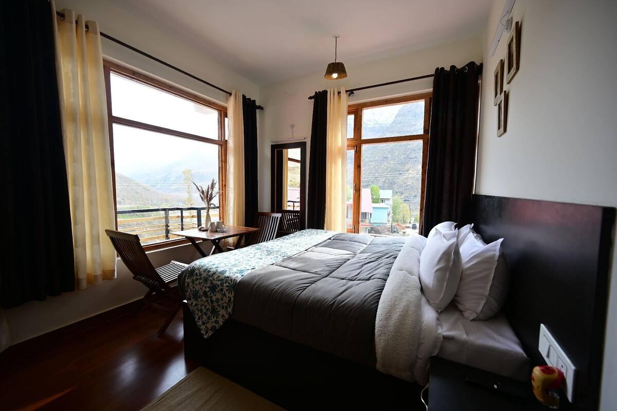 A bedroom with a large bed, a desk, and a chair, a window with a view of mountains, and a ceiling light.
