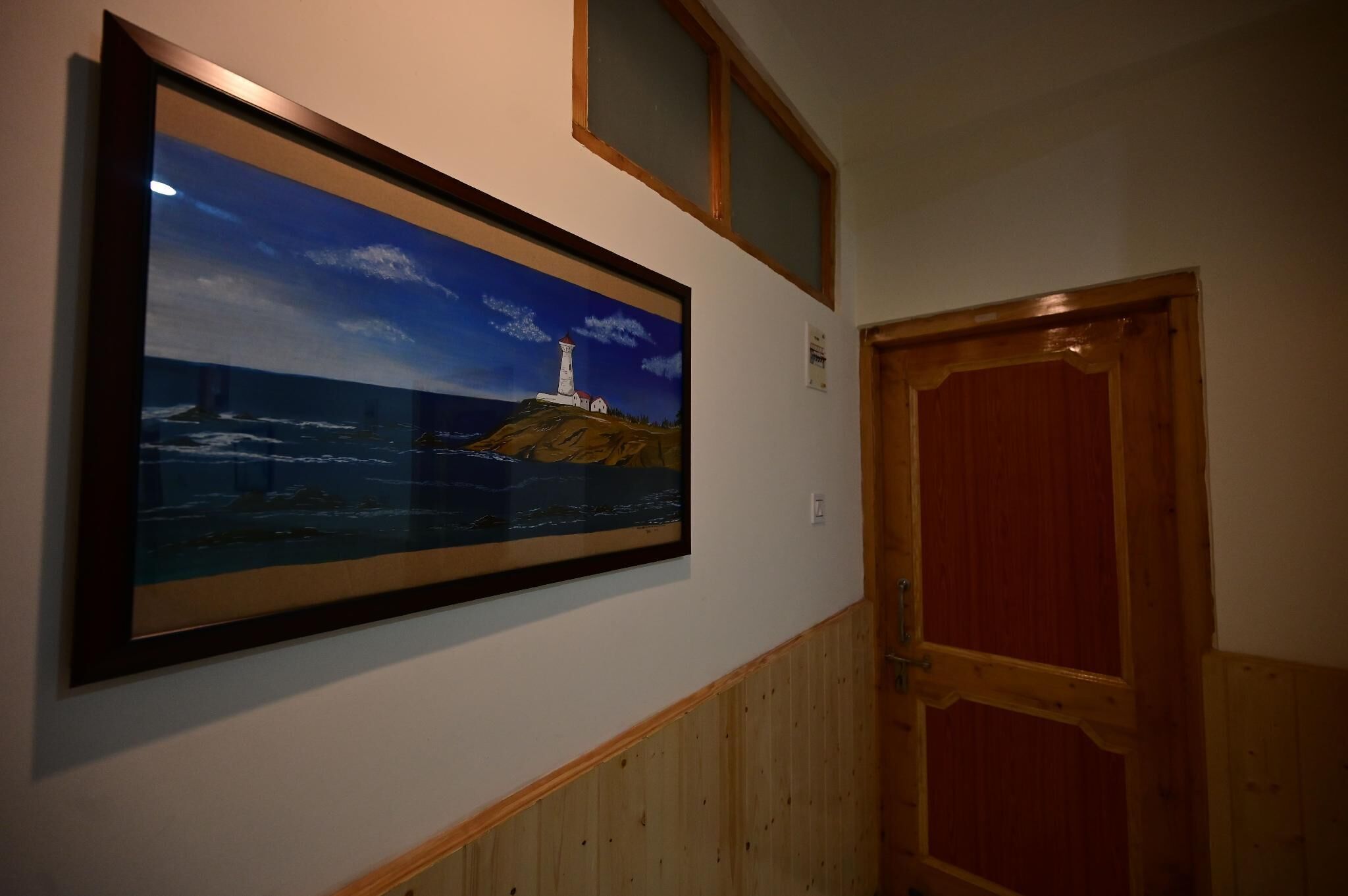 A framed painting of a lighthouse on a cliff, a wooden door, and a window above the door.