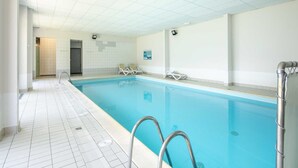 A swimming pool with clear blue water, tiled walls, and a tiled floor. There are two metal pool ladders and a door leading to another room.