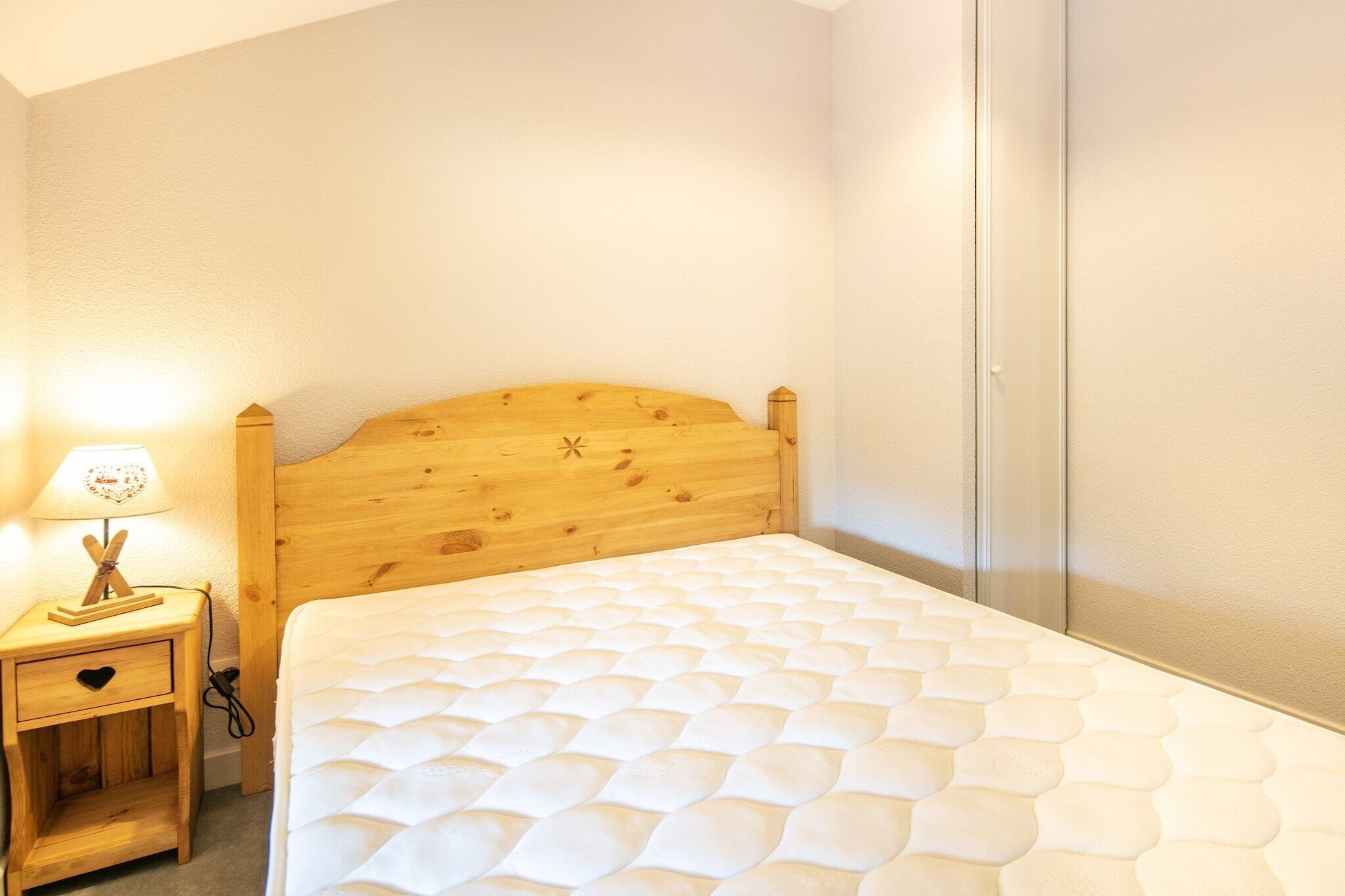 A bedroom with a wooden headboard, a bedside table with a lamp, and a white mattress.