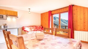 A dining area with a table set for two, wooden chairs, a red patterned sofa, a microwave, and a window with a view of mountains.