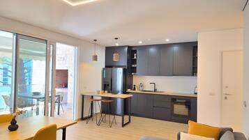 A modern kitchen with dark cabinets, a built-in oven, and a sink. There is a dining area with a table and chairs, and a sofa with cushions.