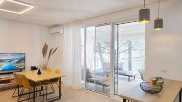A modern living room with a dining area, a TV displaying a scenic image, and a large glass door leading to an outdoor seating area.