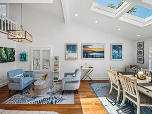 A modern living room with a dining area, featuring a skylight, artwork, and a mix of furniture styles.
