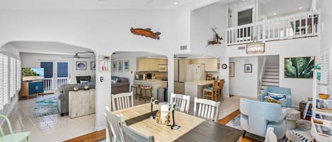 A spacious living area with a large wooden dining table, white chairs, and a kitchen in the background.