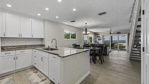 A spacious kitchen with white cabinets, a granite countertop, and a large island. There is a dining area with a table and chairs, and a view of the ocean through large sliding glass doors.