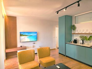 A modern living room with a flat-screen TV, two yellow armchairs, a wooden bench, a kitchen area with teal cabinets, and a wooden floor. - SanPietro Holiday Homes 296 (Gjiri Lalzit)