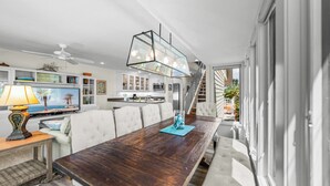 Un vaste espace de vie comprenant une grande table à manger en bois, des chaises à revêtement blanc, un téléviseur et, au fond, une cuisine.