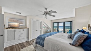 Une chambre à coucher comprenant un lit, un ventilateur de plafond, une télévision, une armoire et une vue sur la mer, visible à travers une grande fenêtre.