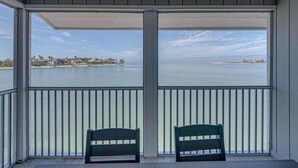 Ein überdachter Balkon mit zwei Stühlen, der Blick auf ein ruhiges Meer und eine entfernte Küstenlinie mit Gebäuden freigibt.