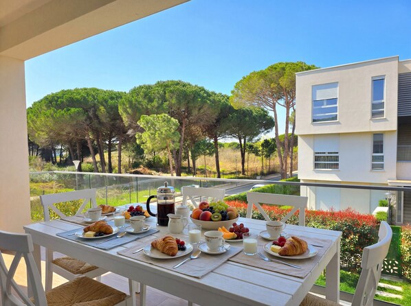 A table set with breakfast items, including croissants, fruit, and beverages, on a balcony with a view of trees and a building.