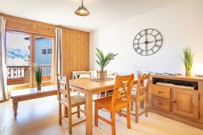 A dining area with a wooden table and chairs, a clock on the wall, and a view of mountains through the window.