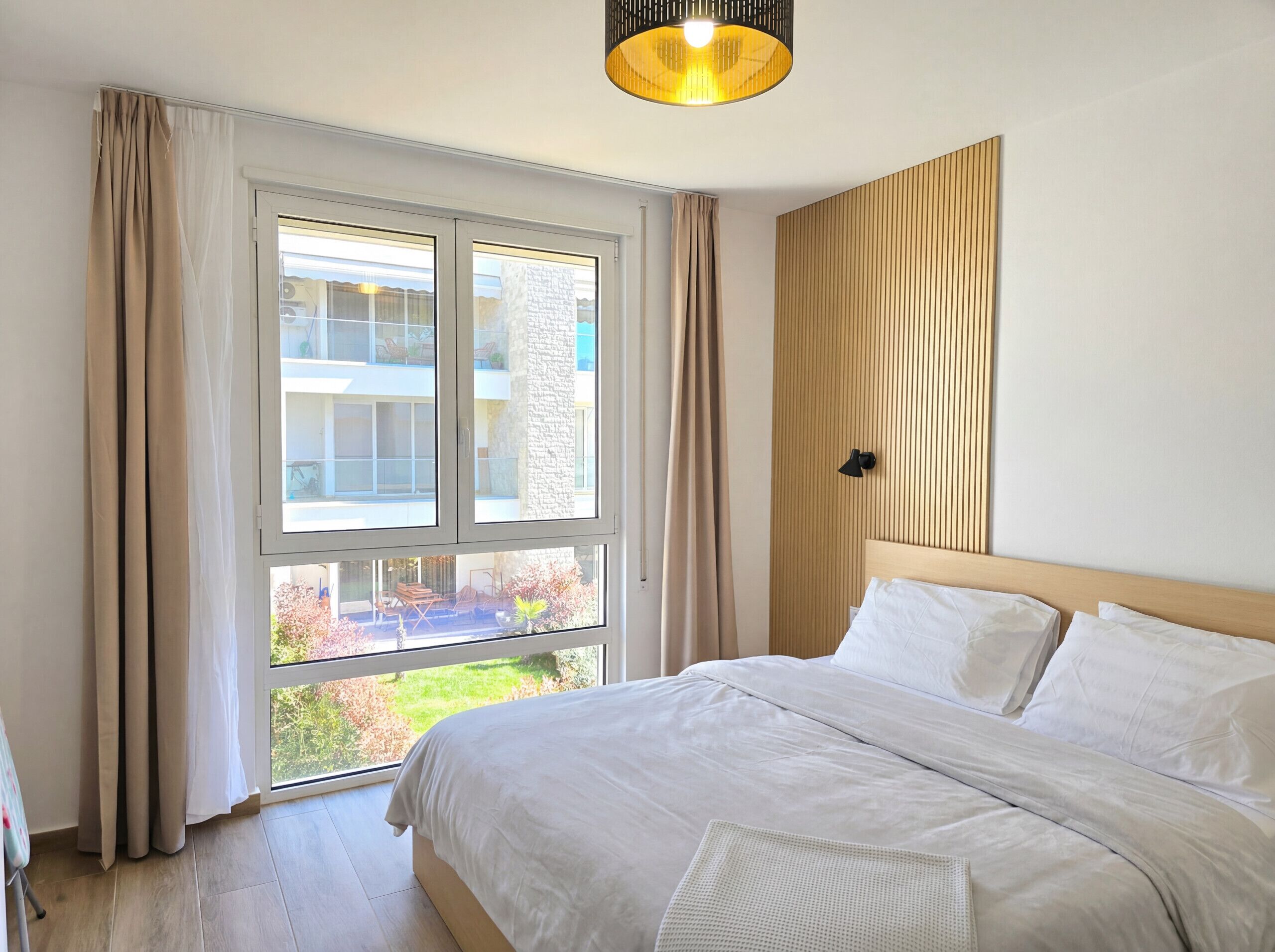 A bedroom with a large window, a bed with white linens, a wooden headboard, and a pendant light.
