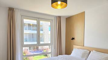 A bedroom with a large window, a bed with white linens, a wooden headboard, and a pendant light.