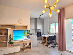 Uma sala moderna com televisão de tela plana, estantes de madeira, área de jantar com mesa e cadeiras, cozinha com armários e uma janela grande com cortinas.
