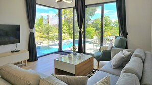 Ein modernes Wohnzimmer mit einem großen Flachbildfernseher, einem Glastisch und einem beigefarbenen Sofa. Durch große Fenster ist ein Blick auf einen Pool und Gartenmöbel möglich.