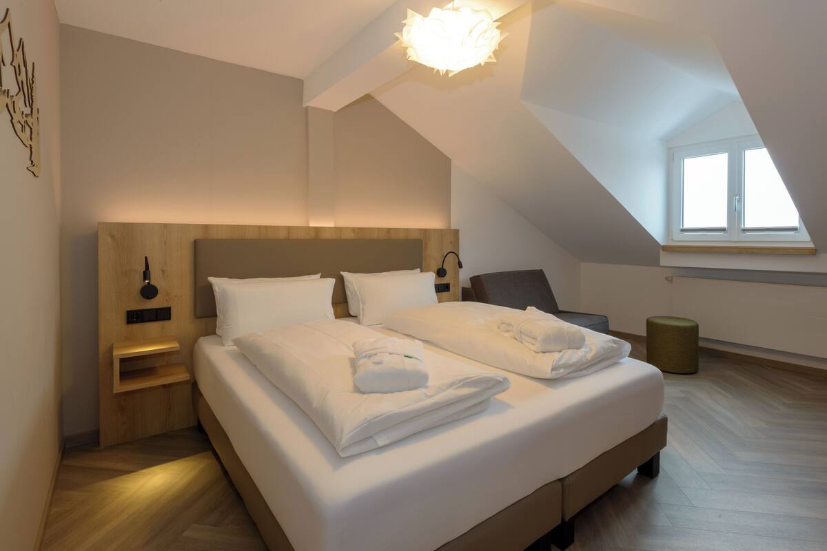 Ein modernes Hotelzimmer mit einem großen Bett, einem Holzkopfstück, einem Nachttisch, einem Sessel, einem kleinen Hocker und einem Oberlicht.