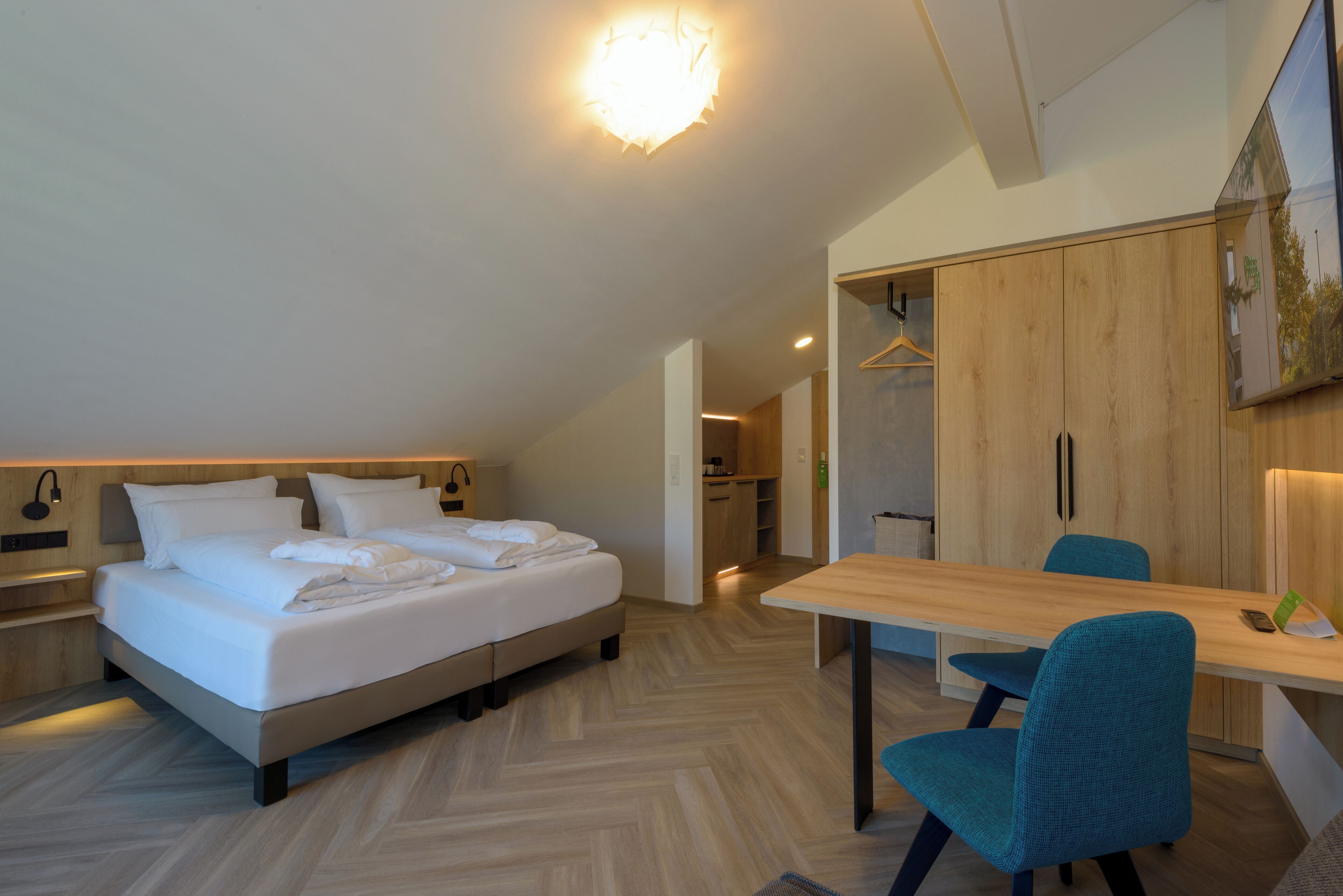 Una habitación de hotel moderna con cama, escritorio y silla. Se observa un armario y un espejo en la pared.