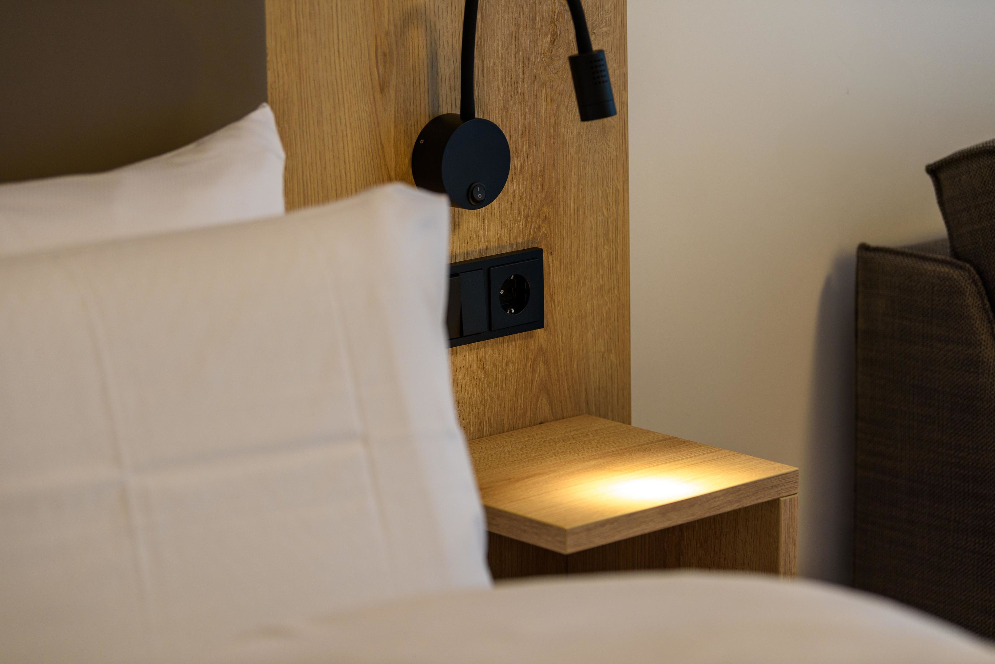 Een net opgemaakt bed met een wit kussen en een houten nachtkastje met een zwarte lamp.