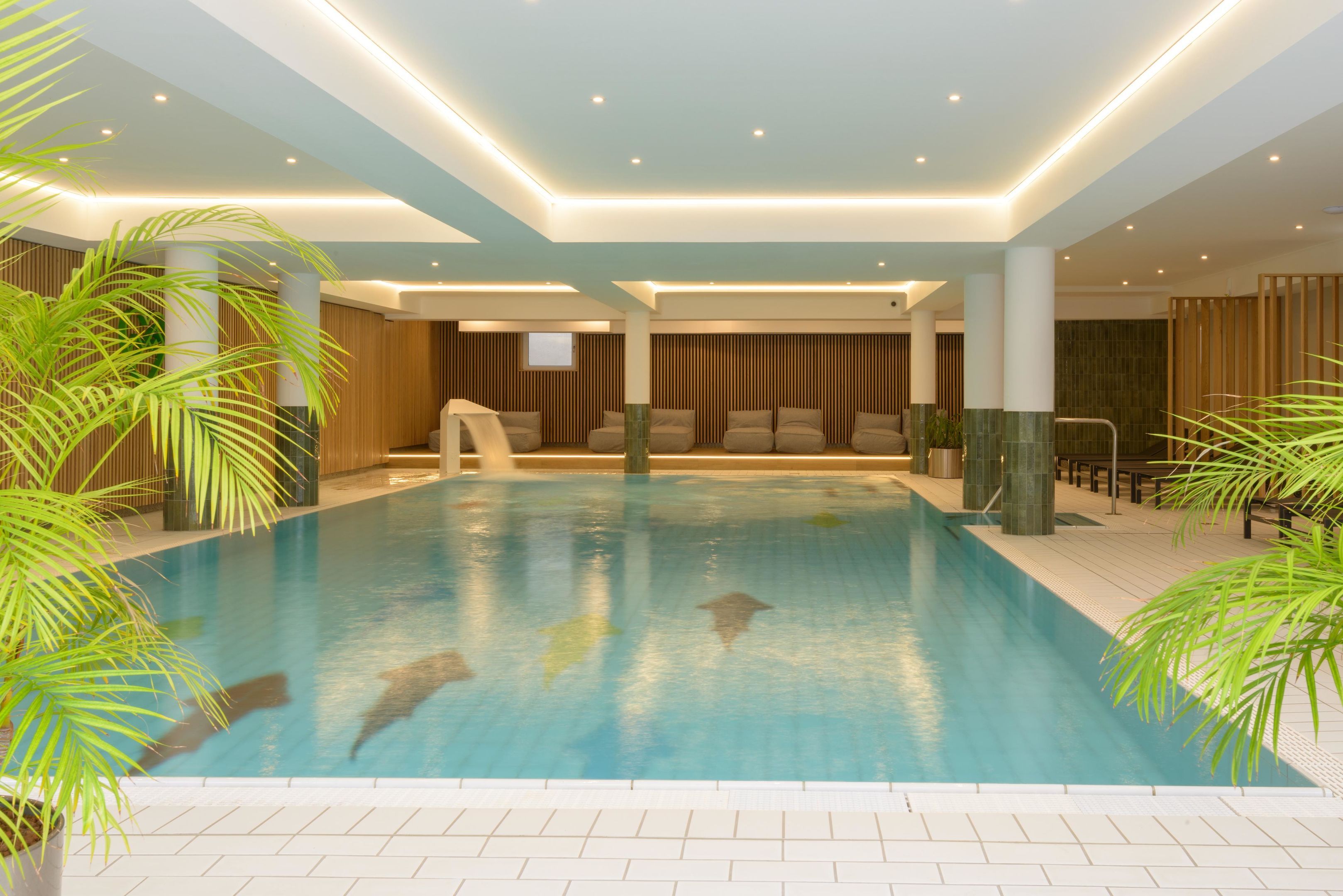 A large indoor swimming pool with multiple lanes, surrounded by seating areas and potted plants.