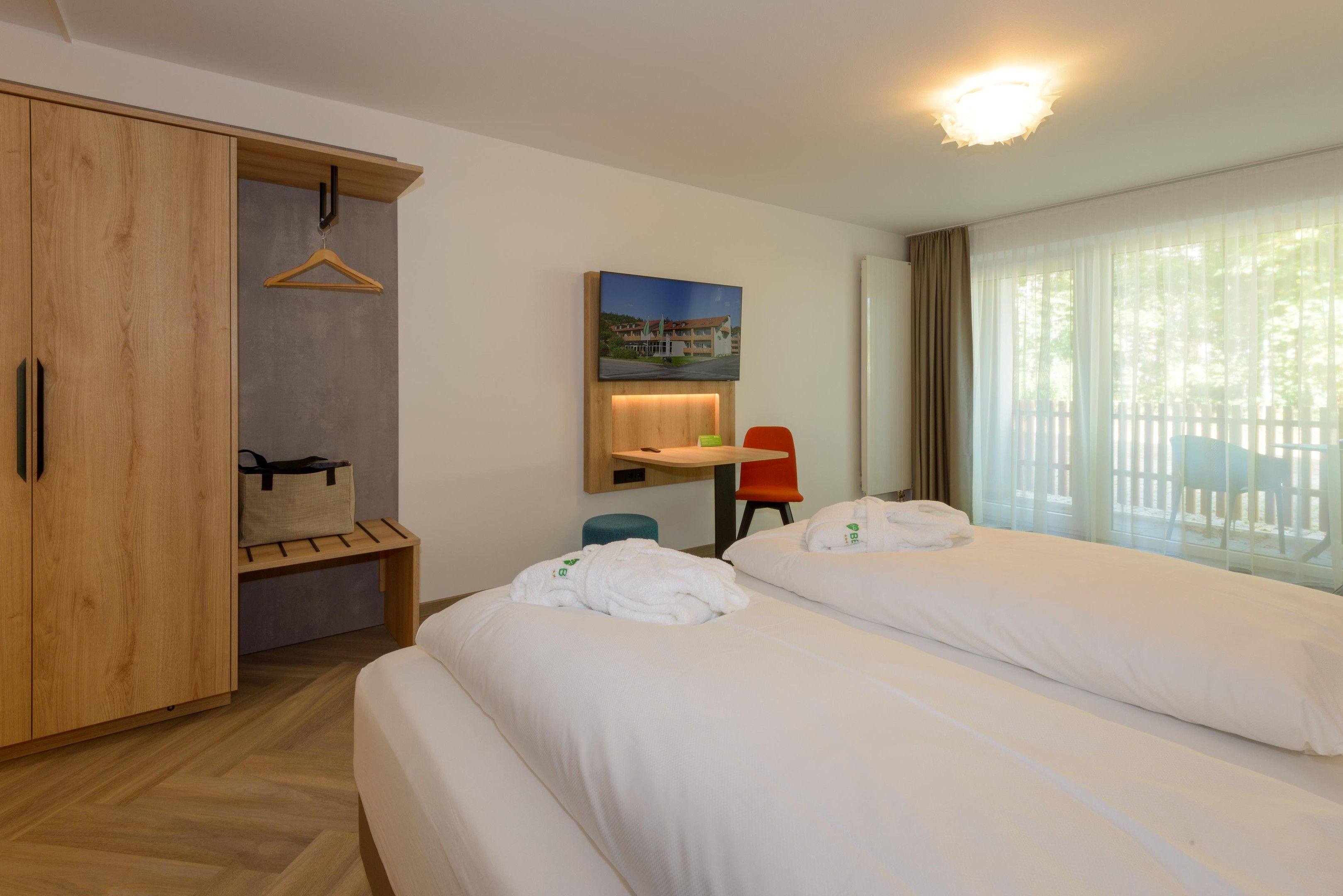 Hotellihuone, jossa on kaksi sänkyä, puinen vaatekaappi, työpöytä tuolilla, naulakko ja ikkuna, jossa on verhot.