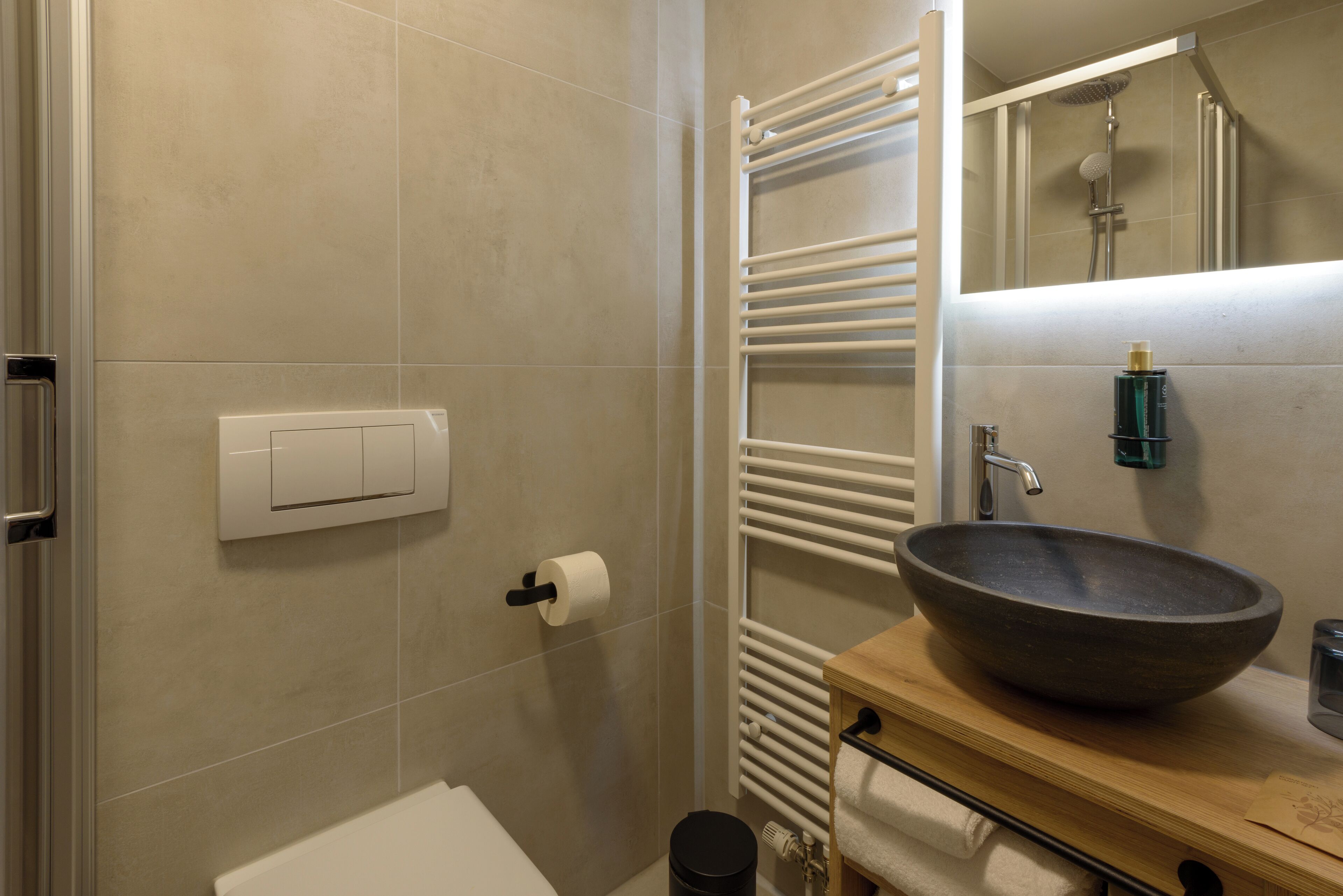 Un baño moderno con pared de azulejos color beige, un inodoro blanco, un tocador de madera con un lavamanos grande y un perchero para toallas, además de un espejo.