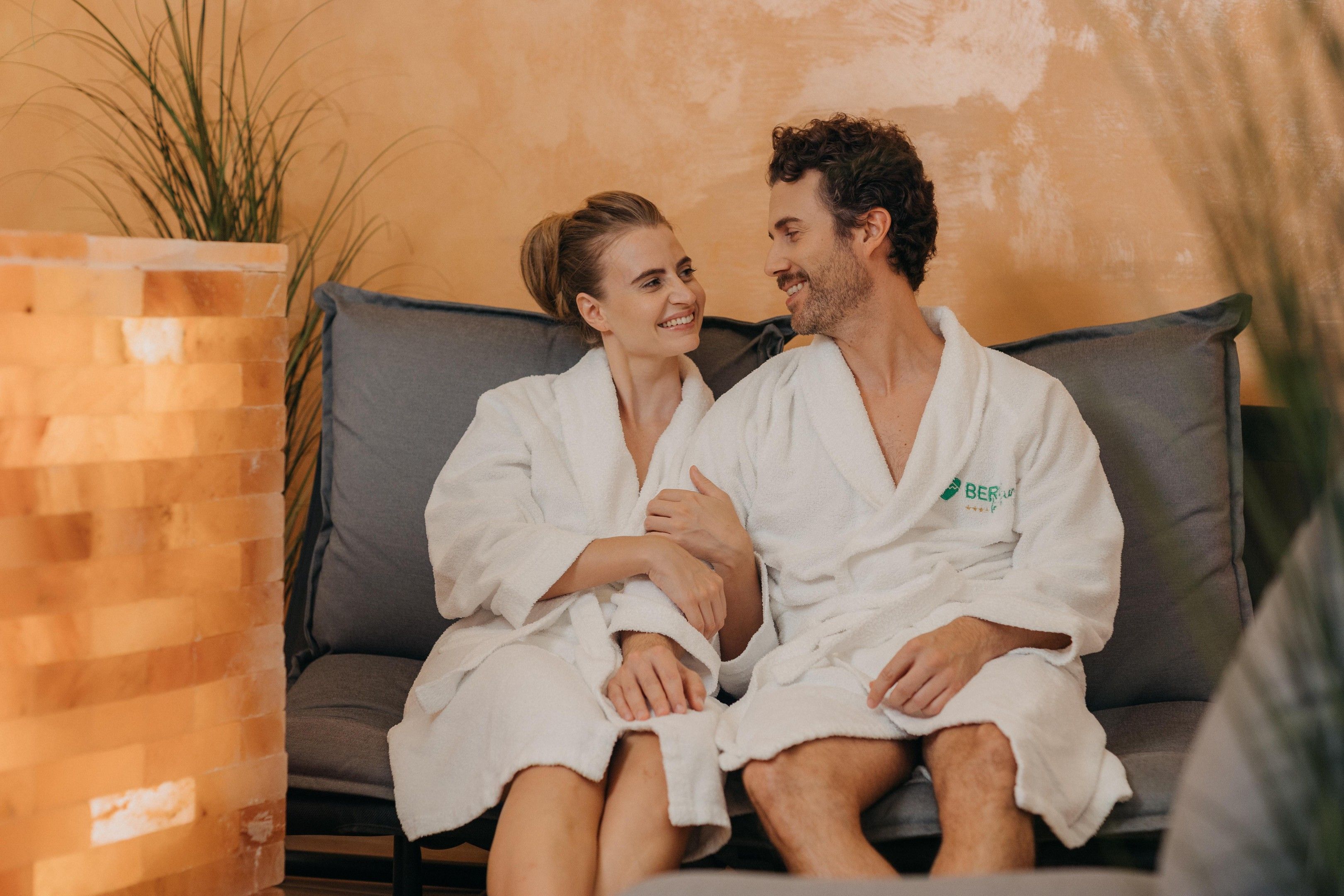 Dos personas con batas blancas sentadas en un sofá gris en una habitación con pared de ladrillo y una planta.