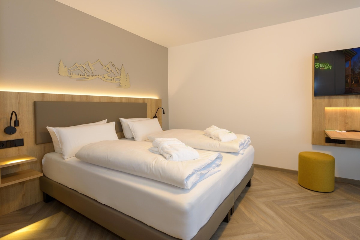 A modern hotel room with a large bed, a wooden headboard, a mounted artwork, a bedside table with a lamp, and a small stool.