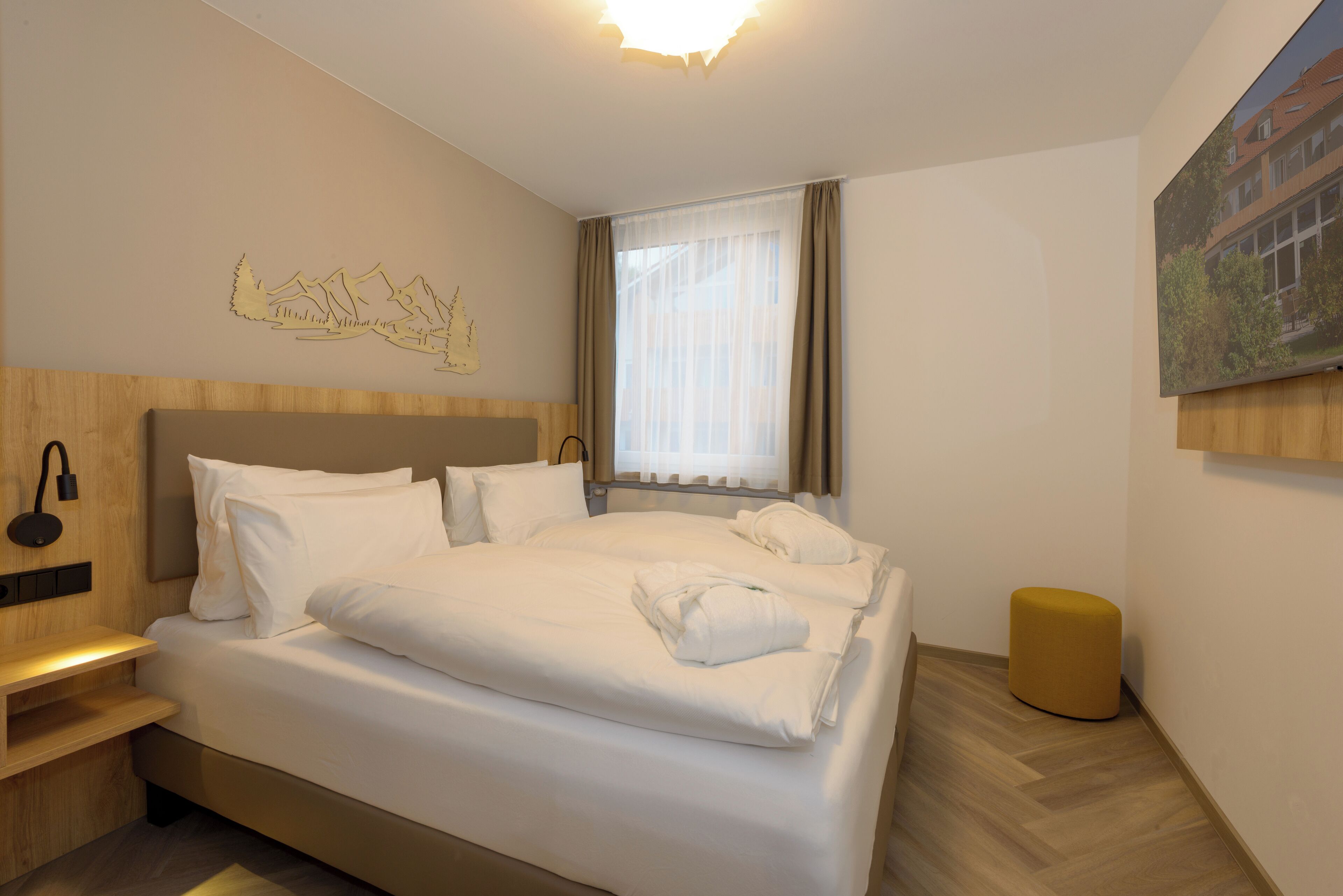 A hotel room with a large bed, a wooden nightstand, a headboard with a light fixture, a window with sheer curtains, and a painting on the wall.