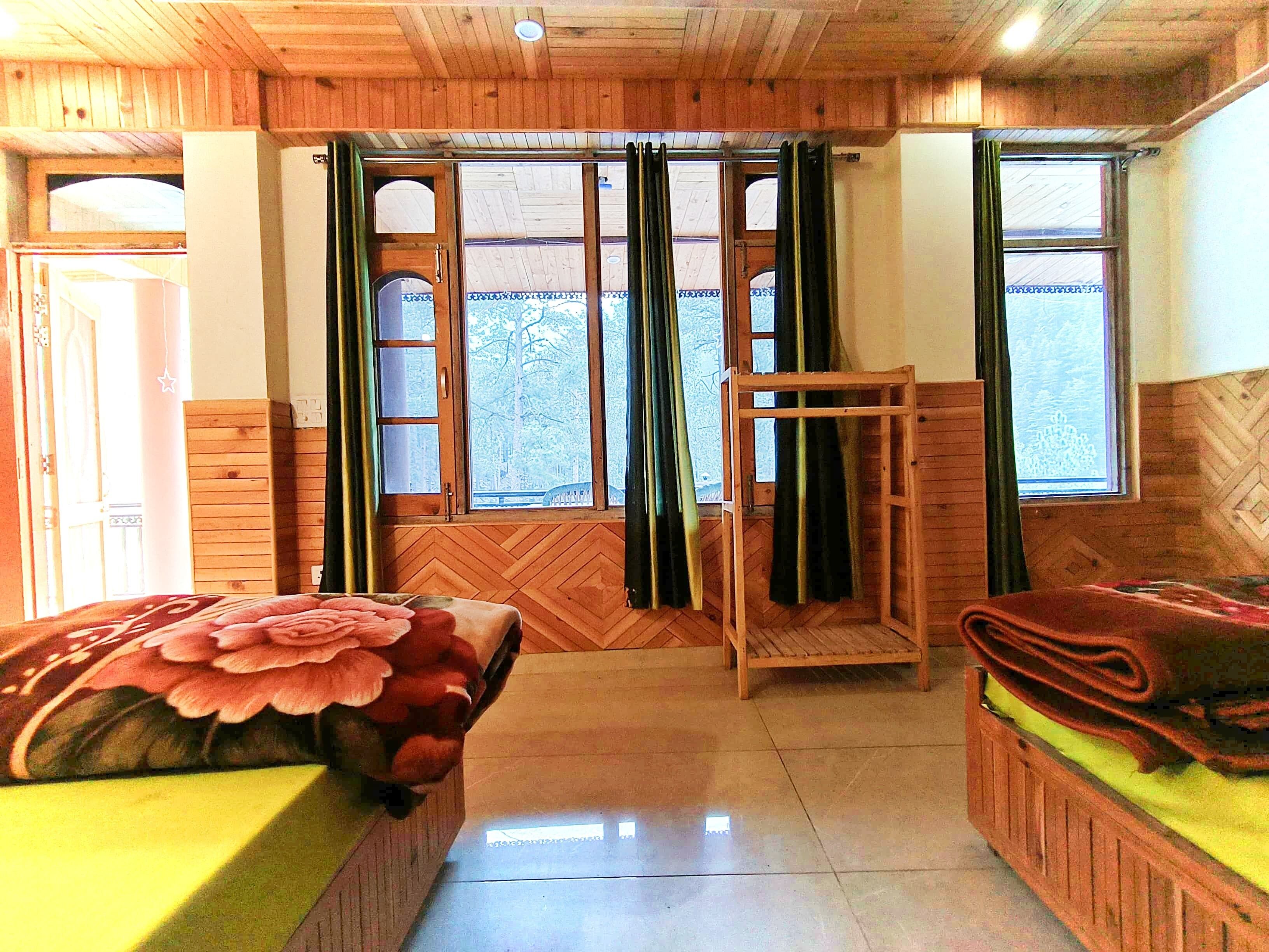 A room with wooden walls and floors, a bed with a floral bedspread, a bench with a green cushion, and a window with curtains.