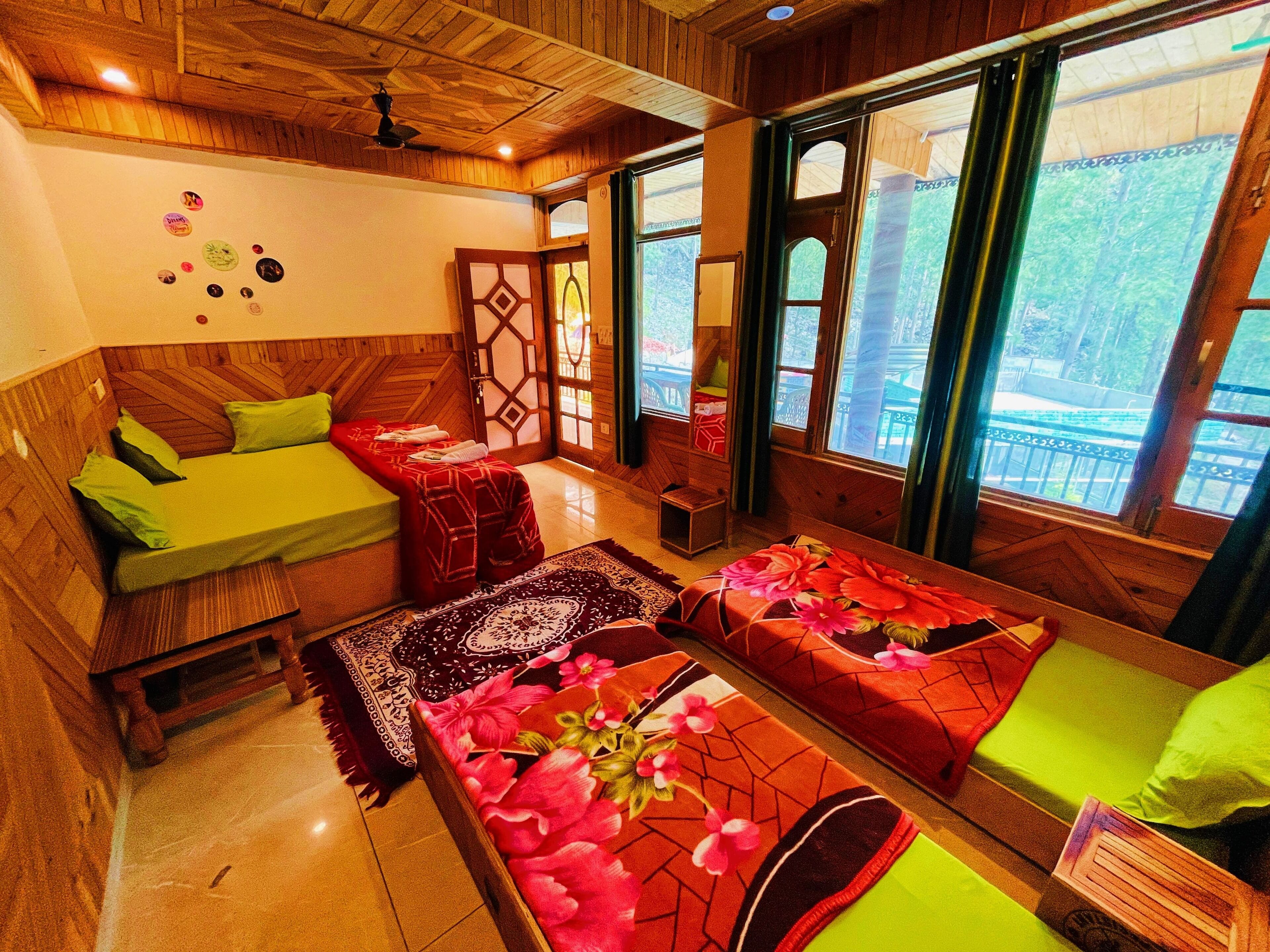 A room with wooden walls and ceiling, a bed with green bedding, a sofa with red and green cushions, and a window with a view of greenery.