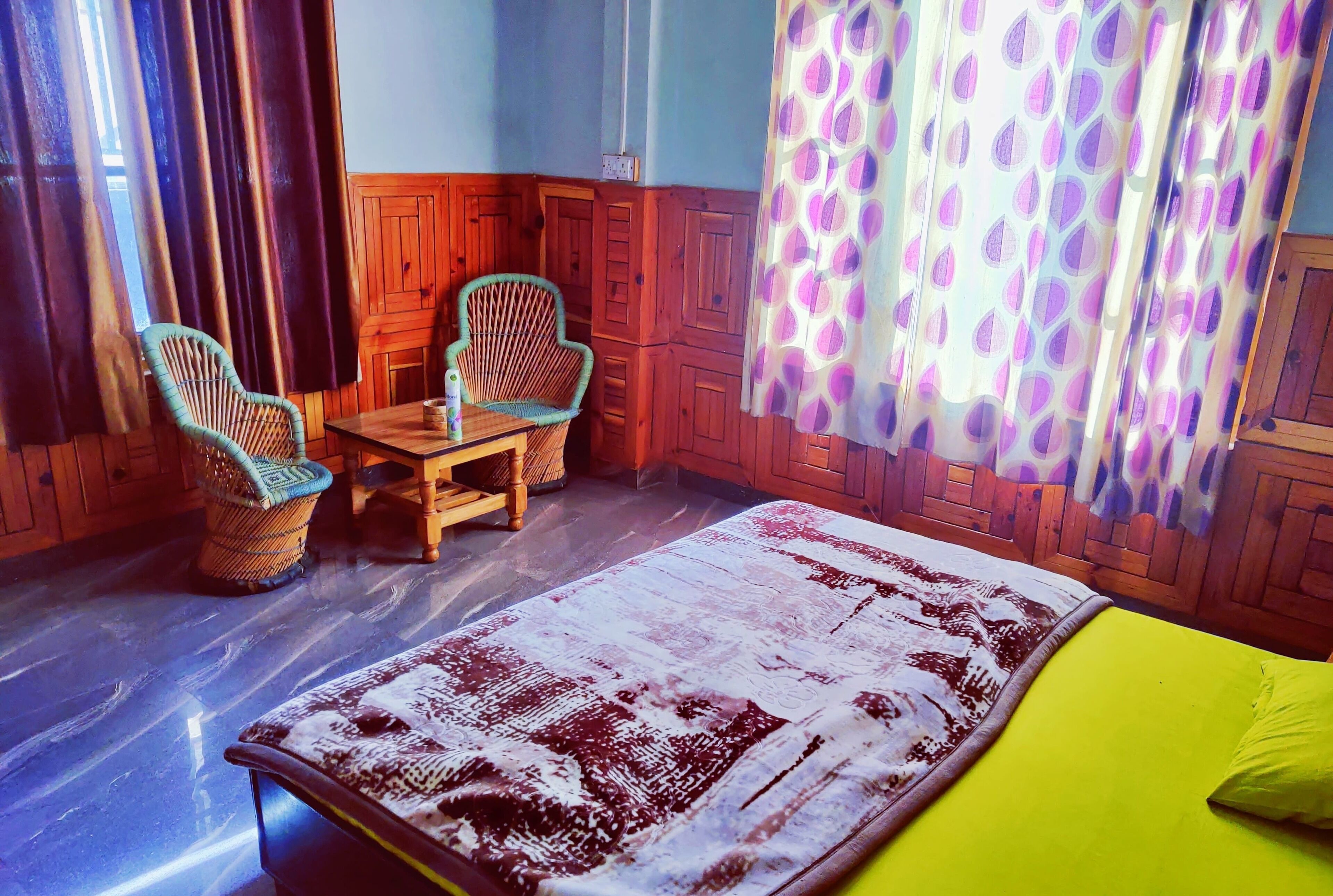 A room with a bed, two wicker chairs, a small table, wooden paneling, and patterned curtains.