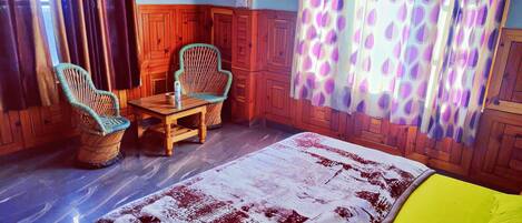 A room with a bed, two wicker chairs, a small table, wooden paneling, and patterned curtains.