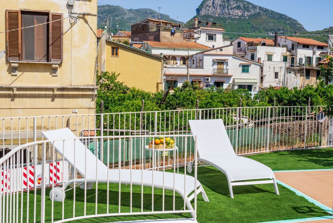 Quartiere residenziale con edifici bianchi, un cortile recintato con due poltrone bianche e una montagna sullo sfondo.