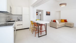 A modern kitchen with white cabinetry, a dining area with a white table and colorful chairs, and a living area with a beige sofa and a cityscape painting.