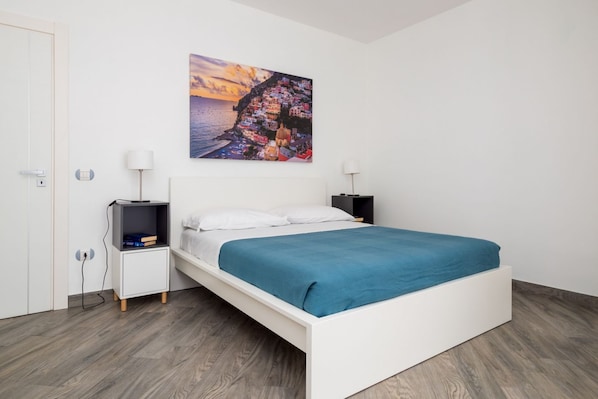 Una camera da letto con un letto bianco, una coperta blu, un comodino con una lampada, un quadro appeso al muro e una porta.