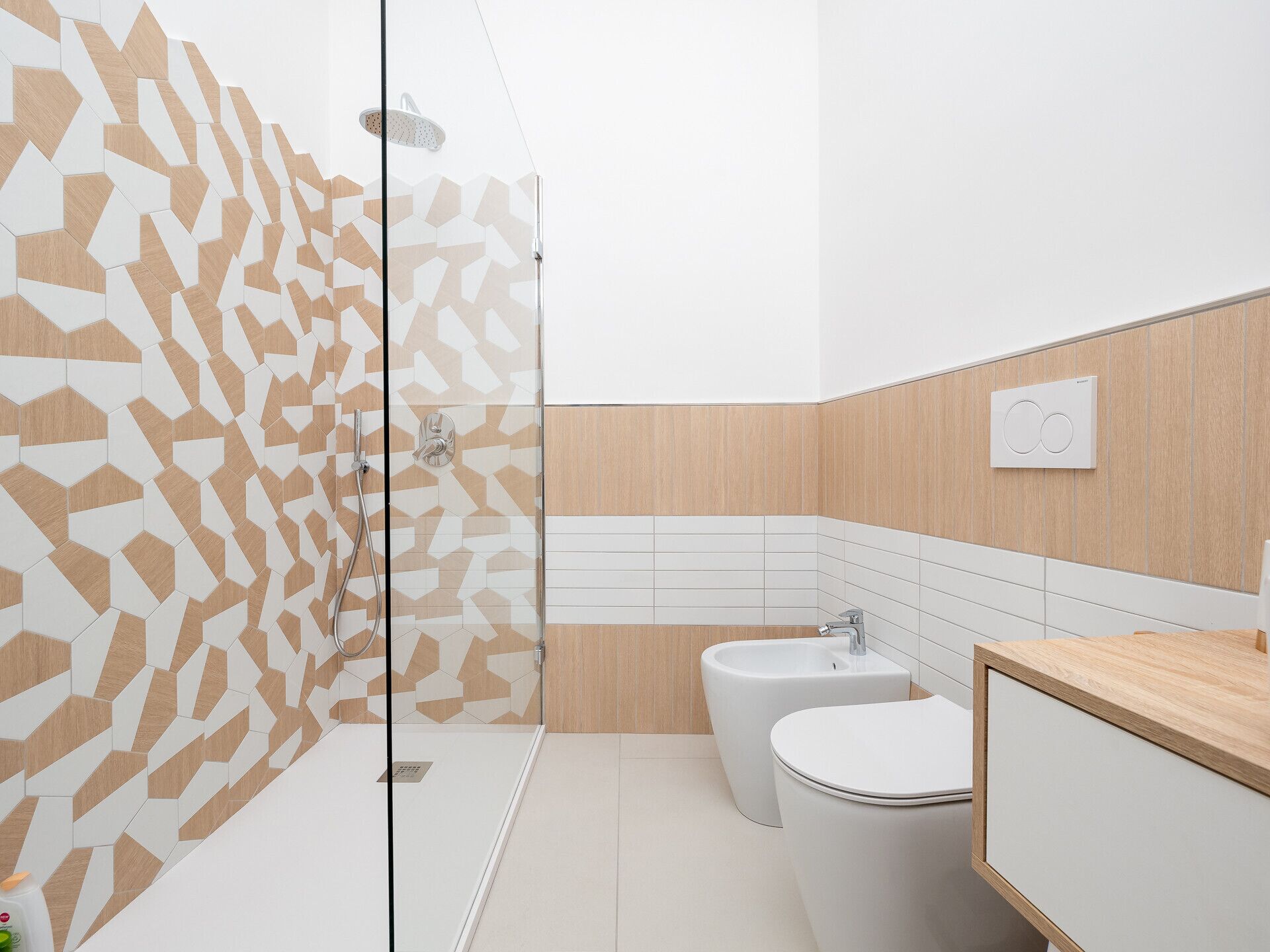 Une salle de bain moderne avec un mur recouvert de carreaux aux motifs géométriques, une douche en verre, un bidet et un meuble-lavabo en bois.