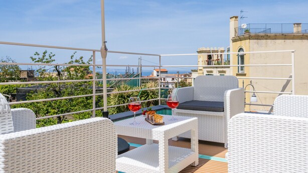 Una terraza en la azotea con mobiliario de mimbre blanco, una mesa con copas de vino y una bandeja de comida, y vistas a la ciudad y al mar.