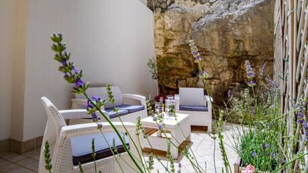 Un patio con mobiliario de mimbre blanco, un muro de piedra y flores moradas.
