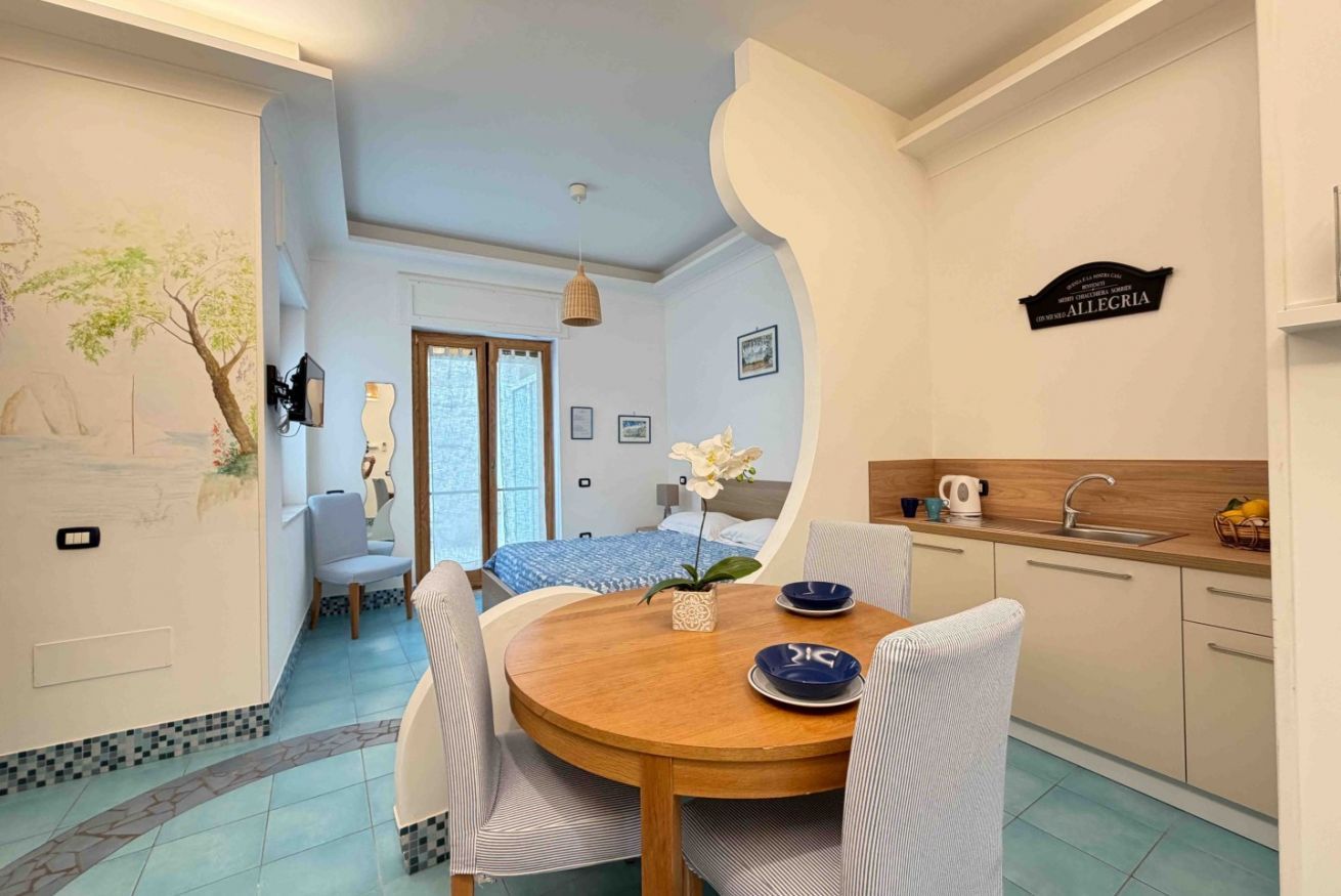 A room with a dining table, chairs, a bed, a kitchen area, and a wall with a sign.