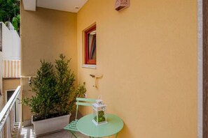 A balcony with a potted plant, a small table with a white lantern, and a window with a red frame.