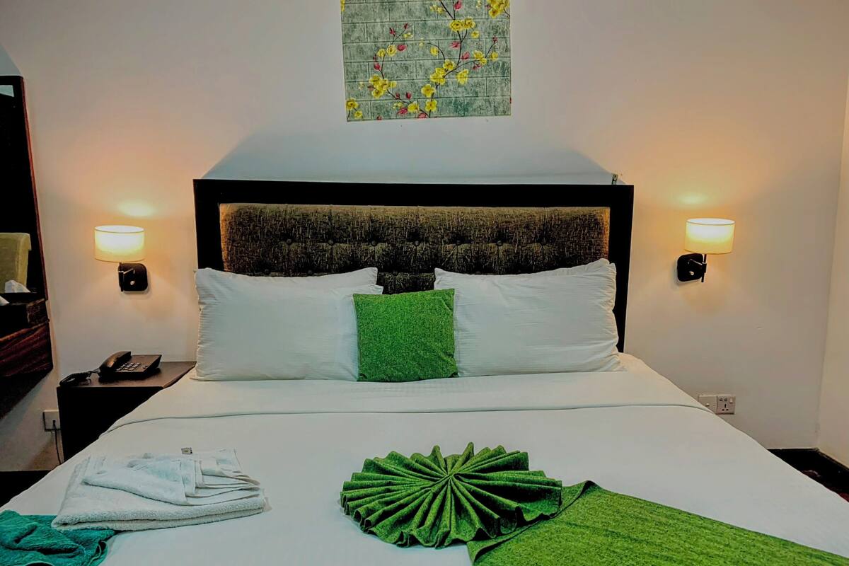 A neatly made bed with white and green pillows, a green throw, and a folded towel on a white bedspread. There are two wall-mounted lamps on either side of the bed.