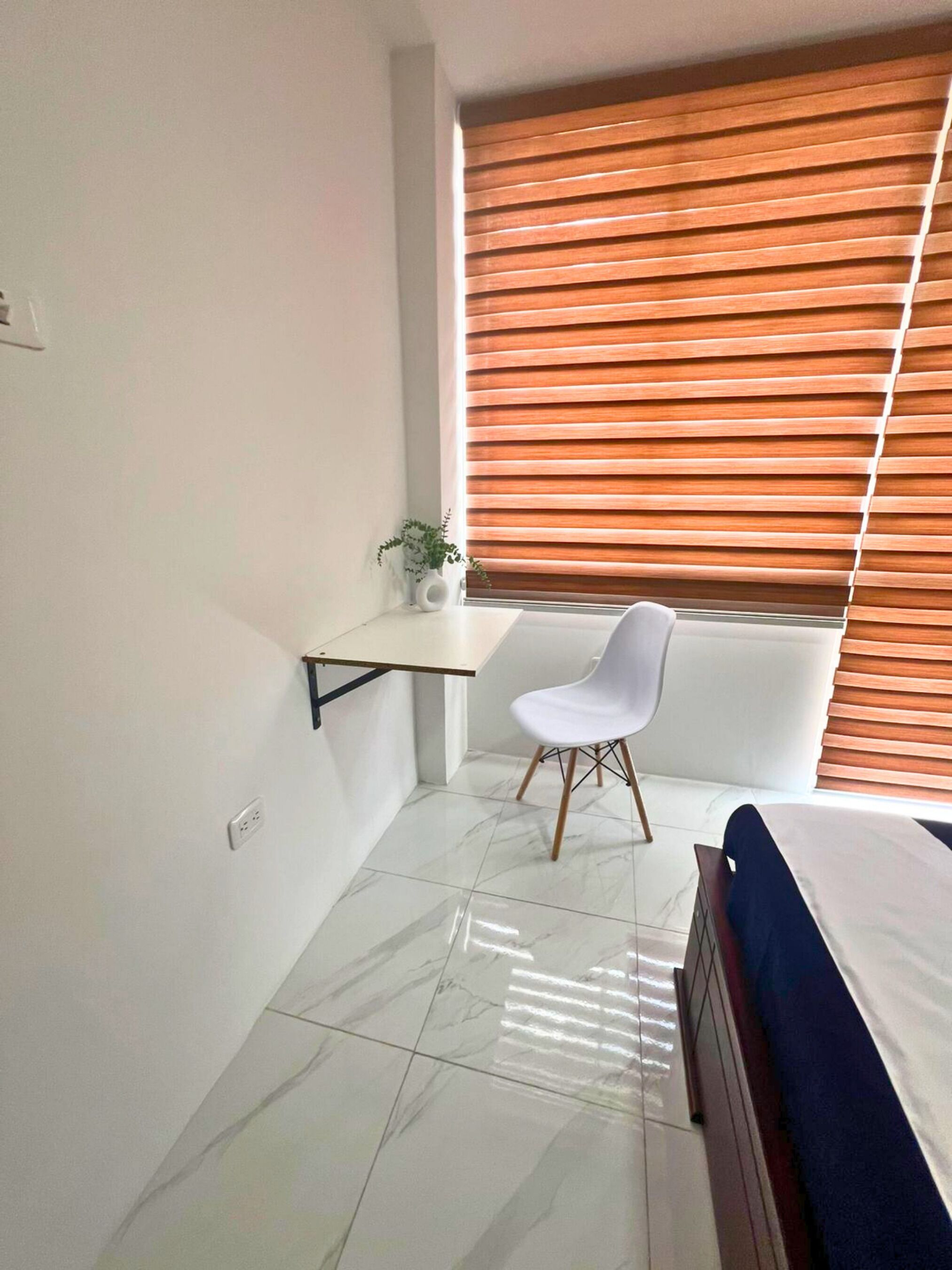 Een slaapkamer met een bed, een witte stoel, een klein plankje en een raam met zonwering.