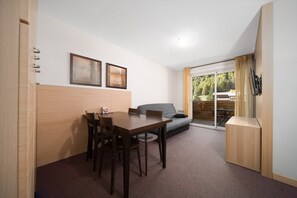 Cozy living space with a dining area, sofa, and flat-screen TV, leading to an outdoor area with a deck and forest views.