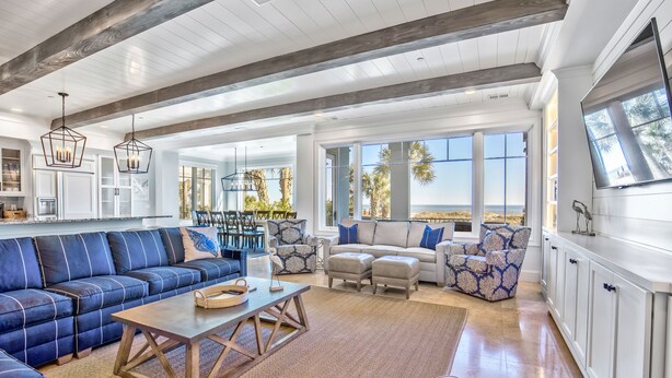 A spacious living area with a large sectional sofa, a coffee table, and patterned armchairs. There are large windows offering a view of palm trees and the ocean. - Oceanfront Estate - Pool, Hot Tub, Pool Table, Bonus Boat- Exclusive Location (Hilton Head Island)