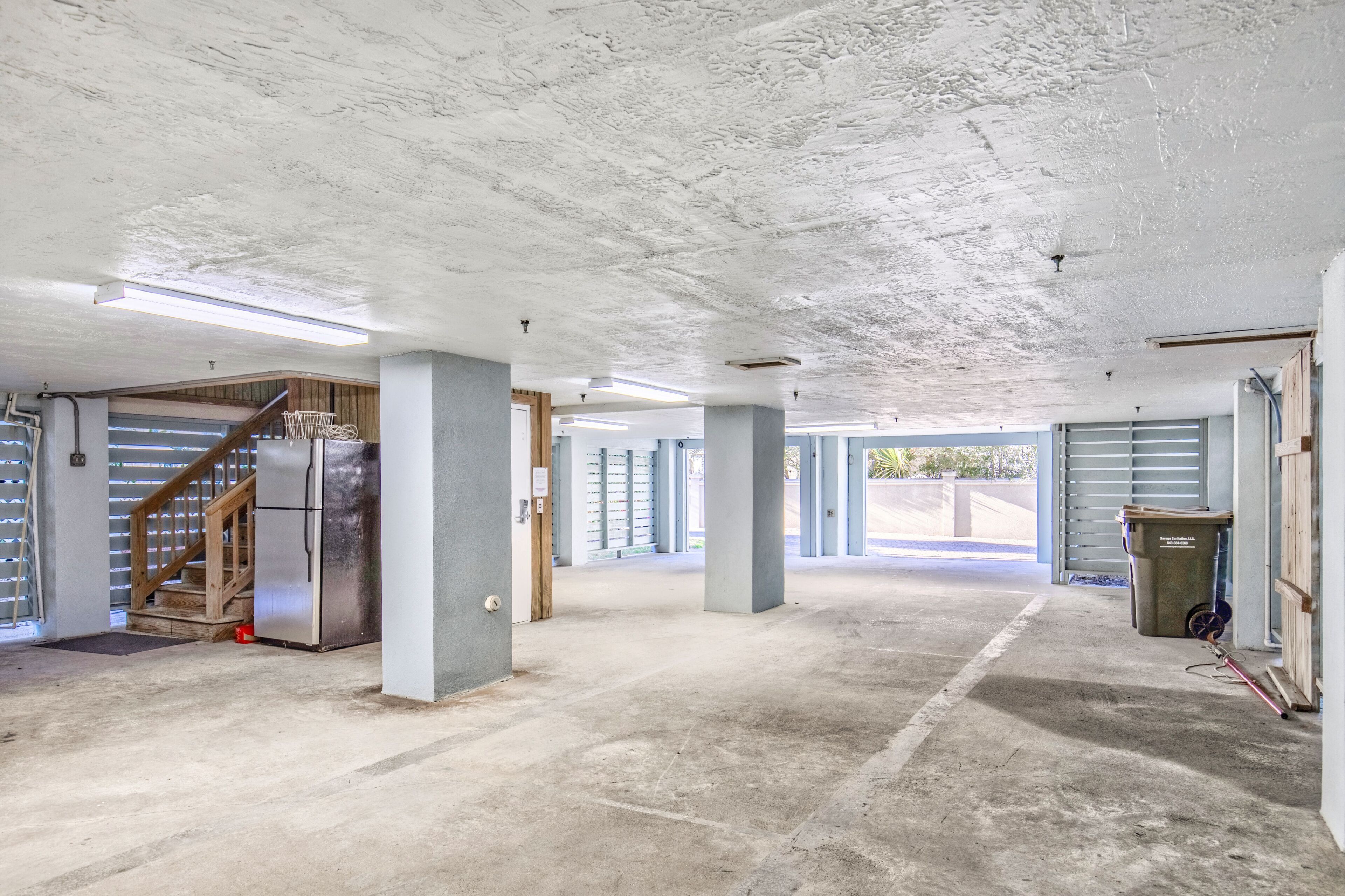A spacious indoor area with concrete floors and white ceilings, featuring a staircase, a refrigerator, and a trash bin.
